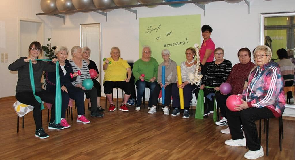 Eine Kursgruppe bei Rolling Pritschen: Frauen sitzen mit Bändern, Bällen und Hanteln nebeneinander und schauen freundlich in die Kamera